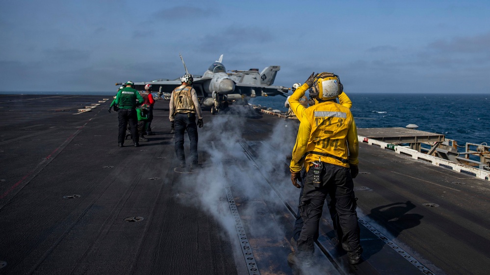 The image shows flight deck operations aboard a naval aircraft carrier at sea. Several crew members in color-coded jerseys are positioned on the flight deck near a military jet aircraft, likely during launch or recovery operations. Steam or exhaust is visible rising from the deck, consistent with catapult launch activity.