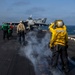 The image shows flight deck operations aboard a naval aircraft carrier at sea. Several crew members in color-coded jerseys are positioned on the flight deck near a military jet aircraft, likely during launch or recovery operations. Steam or exhaust is visible rising from the deck, consistent with catapult launch activity.