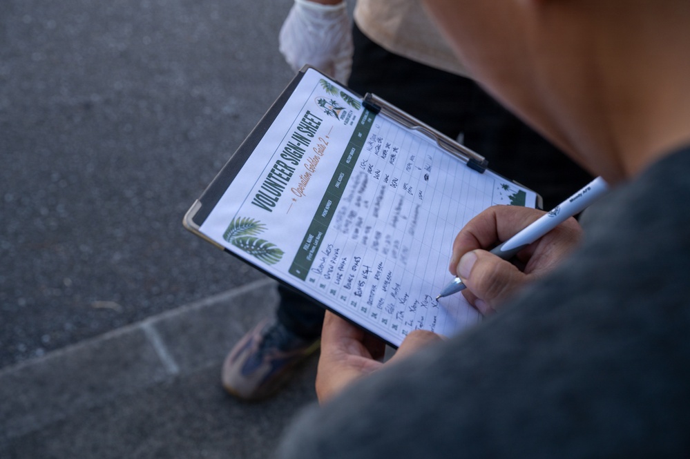A person is holding a clipboard and writing on a 'Volunteer Sign-In Sheet' with a pen. The sheet appears to be associated with a community garden or environmental initiative, featuring green plant/palm leaf graphics. Several names and entries have already been filled in on the lined form.