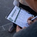 A person is holding a clipboard and writing on a 'Volunteer Sign-In Sheet' with a pen. The sheet appears to be associated with a community garden or environmental initiative, featuring green plant/palm leaf graphics. Several names and entries have already been filled in on the lined form.