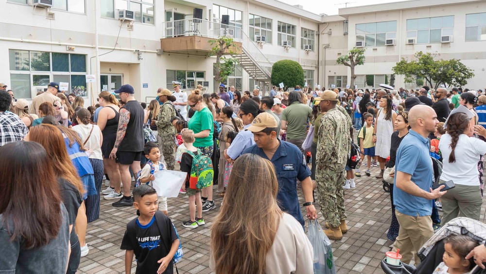 A large crowd of adults and children gathered in an outdoor courtyard of a multi-story institutional building. Several individuals are wearing U.S. Navy military uniforms (NWU/camouflage pattern), suggesting this is a military base event, likely a school orientation or back-to-school day. Children with backpacks and families with bags are present, indicating a school-related gathering.