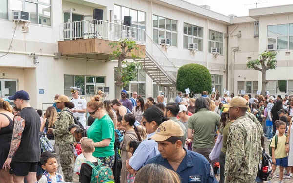 A large crowd of adults and children gathered in an outdoor courtyard of a multi-story institutional building. Several individuals are wearing U.S. Navy military uniforms (NWU/camouflage pattern), suggesting this is a military base event, likely a school orientation or back-to-school day. Children with backpacks and families with bags are present, indicating a school-related gathering.