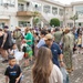 A large crowd of adults and children gathered in an outdoor courtyard of a multi-story institutional building. Several individuals are wearing U.S. Navy military uniforms (NWU/camouflage pattern), suggesting this is a military base event, likely a school orientation or back-to-school day. Children with backpacks and families with bags are present, indicating a school-related gathering.