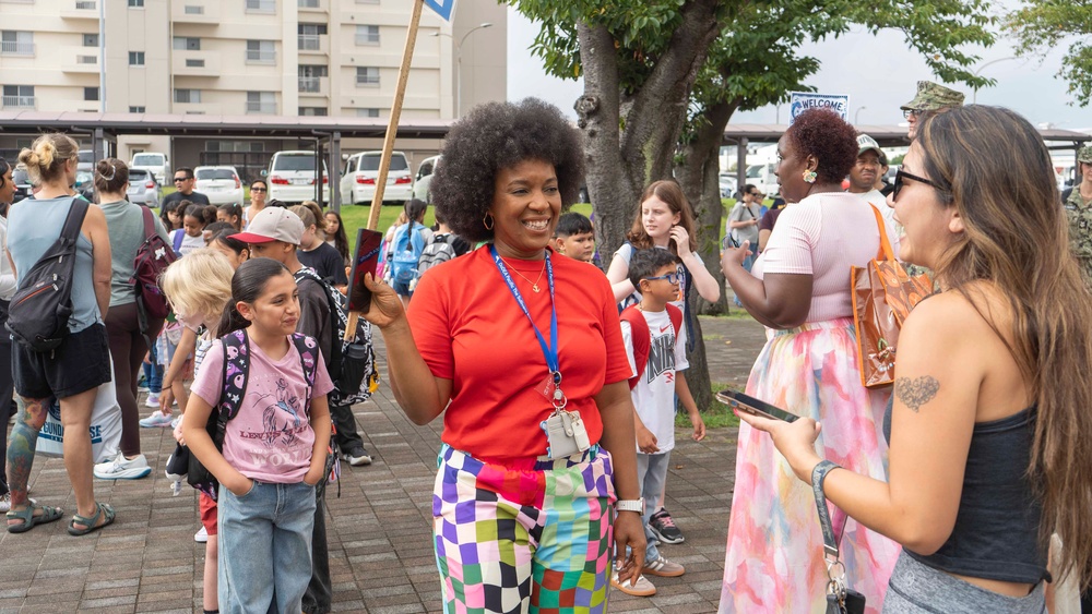 A cheerful woman wearing a red shirt, colorful checkered pants, and a blue lanyard with ID badge stands at the center of an outdoor gathering, smiling and holding a wooden stick or sign. The scene appears to be a school event, likely a first day of school or orientation, with children wearing backpacks and adults gathered in a paved outdoor area. A 'WELCOME' sign is partially visible in the background near what appears to be a military or institutional building complex.
