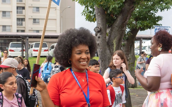 A cheerful woman wearing a red shirt, colorful checkered pants, and a blue lanyard with ID badge stands at the center of an outdoor gathering, smiling and holding a wooden stick or sign. The scene appears to be a school event, likely a first day of school or orientation, with children wearing backpacks and adults gathered in a paved outdoor area. A 'WELCOME' sign is partially visible in the background near what appears to be a military or institutional building complex.