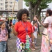 A cheerful woman wearing a red shirt, colorful checkered pants, and a blue lanyard with ID badge stands at the center of an outdoor gathering, smiling and holding a wooden stick or sign. The scene appears to be a school event, likely a first day of school or orientation, with children wearing backpacks and adults gathered in a paved outdoor area. A 'WELCOME' sign is partially visible in the background near what appears to be a military or institutional building complex.