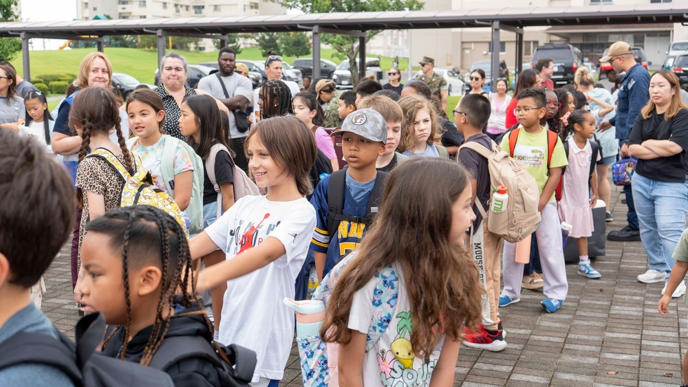 A large group of elementary school-aged children are gathered outdoors in what appears to be a school or community setting, likely during arrival or dismissal time. Adults, possibly parents and school staff, are supervising the children in the background. The setting appears to be on or near a military base, suggested by the presence of individuals in camouflage uniforms and the institutional building architecture.