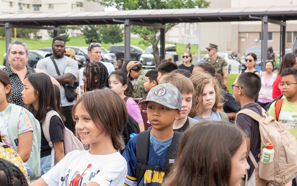 A large group of elementary school-aged children are gathered outdoors in what appears to be a school or community setting, likely during arrival or dismissal time. Adults, possibly parents and school staff, are supervising the children in the background. The setting appears to be on or near a military base, suggested by the presence of individuals in camouflage uniforms and the institutional building architecture.