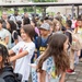 A large group of elementary school-aged children are gathered outdoors in what appears to be a school or community setting, likely during arrival or dismissal time. Adults, possibly parents and school staff, are supervising the children in the background. The setting appears to be on or near a military base, suggested by the presence of individuals in camouflage uniforms and the institutional building architecture.