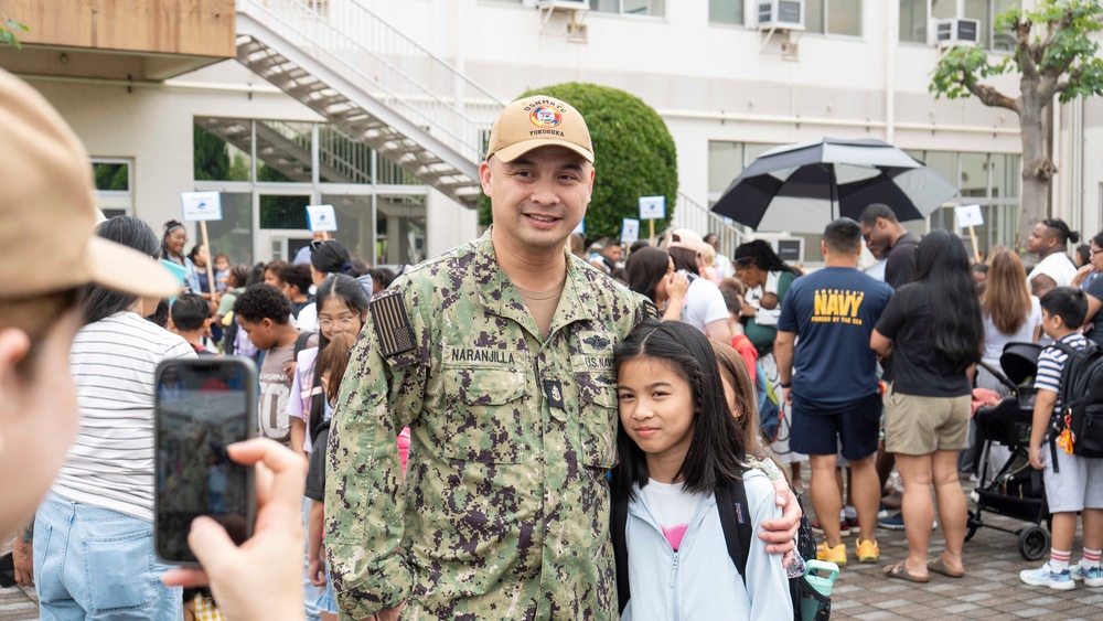A U.S. Navy service member named Naranjilla, wearing NWU Type III camouflage uniform and a cap reading 'Yokosuka', poses with a young girl while someone photographs them. The setting appears to be a school or base facility event, with a large crowd of families and children gathered in the background. Based on the uniform branch tape reading 'U.S. Navy' and the Yokosuka cap, this appears to be a military base school event, likely a first day of school or similar occasion.