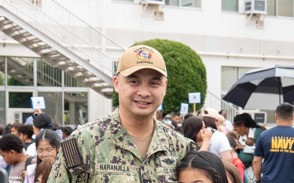 A U.S. Navy service member named Naranjilla, wearing NWU Type III camouflage uniform and a cap reading 'Yokosuka', poses with a young girl while someone photographs them. The setting appears to be a school or base facility event, with a large crowd of families and children gathered in the background. Based on the uniform branch tape reading 'U.S. Navy' and the Yokosuka cap, this appears to be a military base school event, likely a first day of school or similar occasion.