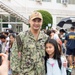 A U.S. Navy service member named Naranjilla, wearing NWU Type III camouflage uniform and a cap reading 'Yokosuka', poses with a young girl while someone photographs them. The setting appears to be a school or base facility event, with a large crowd of families and children gathered in the background. Based on the uniform branch tape reading 'U.S. Navy' and the Yokosuka cap, this appears to be a military base school event, likely a first day of school or similar occasion.
