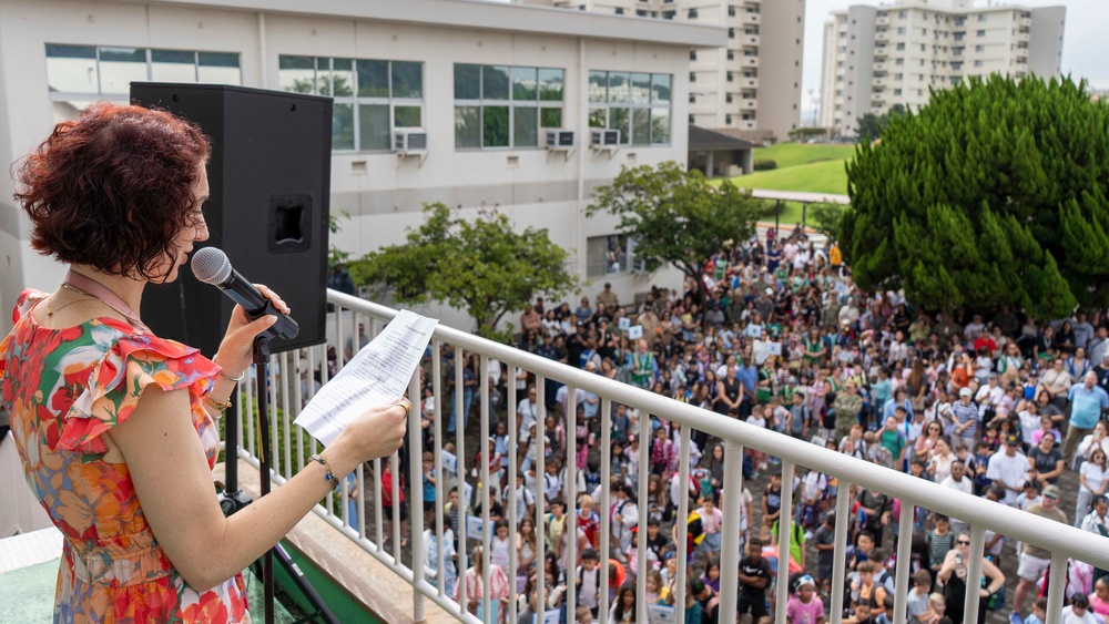 A woman with reddish-brown hair wearing a colorful floral dress stands on an elevated balcony or terrace, speaking into a microphone while holding papers. She is addressing a large crowd of hundreds of people gathered below in an outdoor plaza or courtyard. The setting appears to be a school, community center, or institutional campus based on the surrounding buildings.