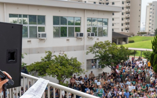A woman with reddish-brown hair wearing a colorful floral dress stands on an elevated balcony or terrace, speaking into a microphone while holding papers. She is addressing a large crowd of hundreds of people gathered below in an outdoor plaza or courtyard. The setting appears to be a school, community center, or institutional campus based on the surrounding buildings.