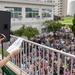 A woman with reddish-brown hair wearing a colorful floral dress stands on an elevated balcony or terrace, speaking into a microphone while holding papers. She is addressing a large crowd of hundreds of people gathered below in an outdoor plaza or courtyard. The setting appears to be a school, community center, or institutional campus based on the surrounding buildings.
