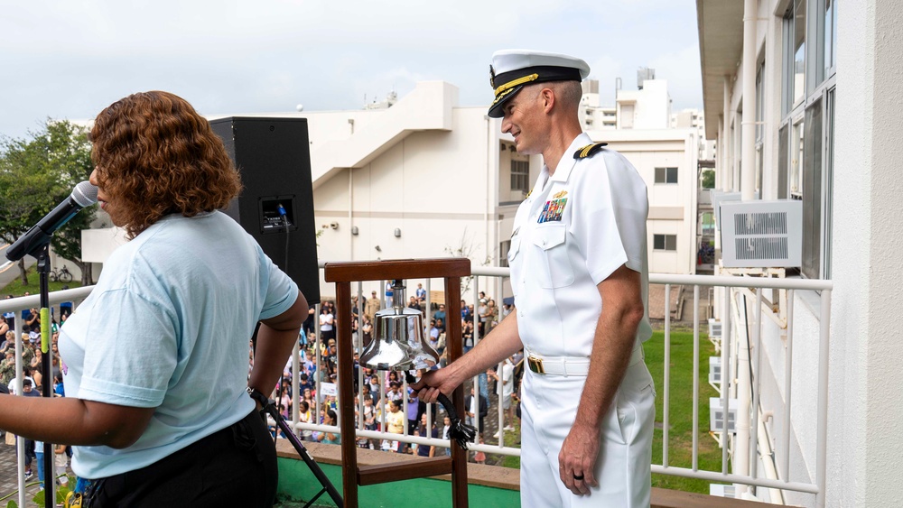 A U.S. Navy officer in white dress uniform stands on an elevated balcony or stage next to a ship's bell mounted on a wooden stand, smiling toward a large crowd gathered below. A woman in a light blue shirt stands at a microphone nearby, suggesting she is speaking or emceeing the event. The setting appears to be an outdoor ceremony at a military installation, likely a school or community event based on the crowd composition.