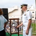 A U.S. Navy officer in white dress uniform stands on an elevated balcony or stage next to a ship's bell mounted on a wooden stand, smiling toward a large crowd gathered below. A woman in a light blue shirt stands at a microphone nearby, suggesting she is speaking or emceeing the event. The setting appears to be an outdoor ceremony at a military installation, likely a school or community event based on the crowd composition.