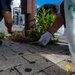 Two individuals are crouching on a brick sidewalk performing what appears to be weeding or cleanup work around the base of a metal pole in an urban setting. One person is wearing white work gloves and holding a small tool, while both carry plastic bags likely for collecting debris. The scene appears to take place in a Japanese city based on visible signage and architectural elements in the background.