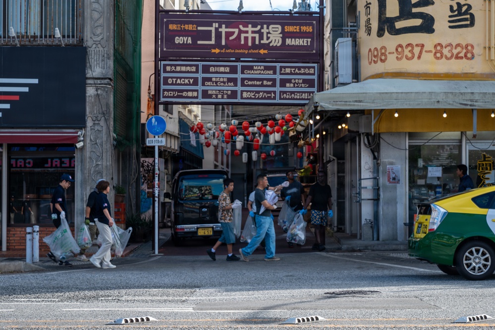 A street scene in front of Goya Market (ゴヤ市場), established in 1955, located in what appears to be Okinawa, Japan. Several people are crossing or walking along the street carrying plastic bags, suggesting a cleanup activity or shopping. The market entrance is decorated with red and white paper lanterns, and a yellow-green taxi is visible on the right side.