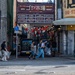 A street scene in front of Goya Market (ゴヤ市場), established in 1955, located in what appears to be Okinawa, Japan. Several people are crossing or walking along the street carrying plastic bags, suggesting a cleanup activity or shopping. The market entrance is decorated with red and white paper lanterns, and a yellow-green taxi is visible on the right side.
