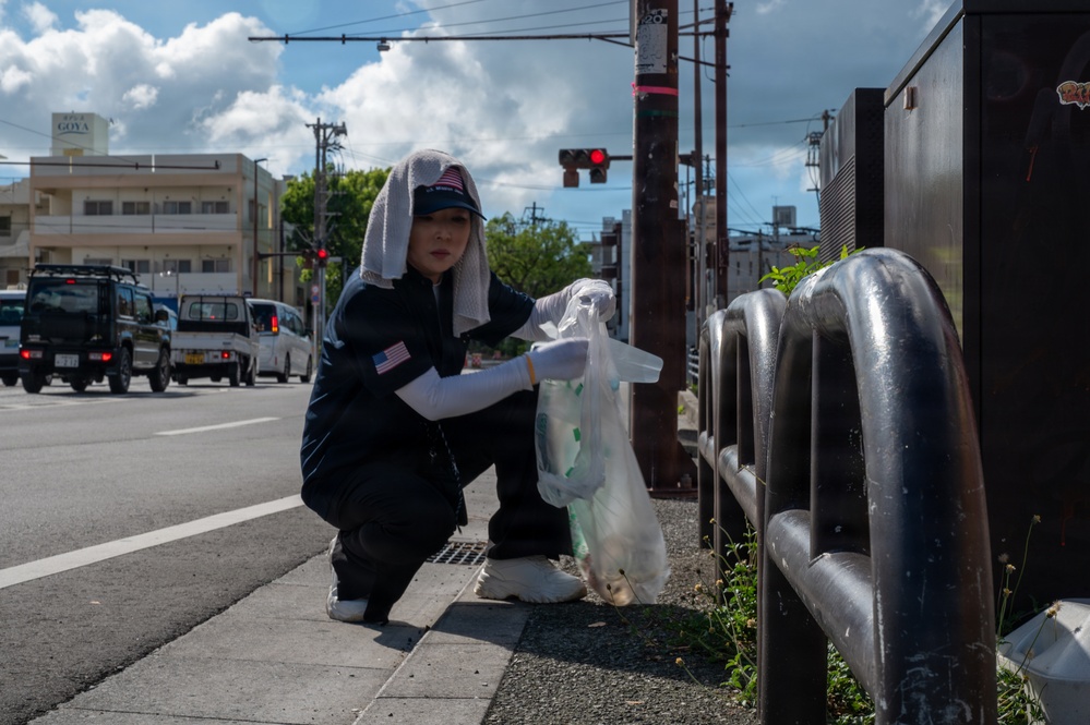 A woman is crouching on a sidewalk near a busy urban intersection, collecting litter into a clear plastic bag. She is wearing a dark uniform with a US flag patch, white gloves, arm sleeves, and has a towel draped over her head under a cap for sun protection. The setting appears to be a Japanese city street, evidenced by Japanese signage and license plates.