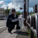 A woman is crouching on a sidewalk near a busy urban intersection, collecting litter into a clear plastic bag. She is wearing a dark uniform with a US flag patch, white gloves, arm sleeves, and has a towel draped over her head under a cap for sun protection. The setting appears to be a Japanese city street, evidenced by Japanese signage and license plates.