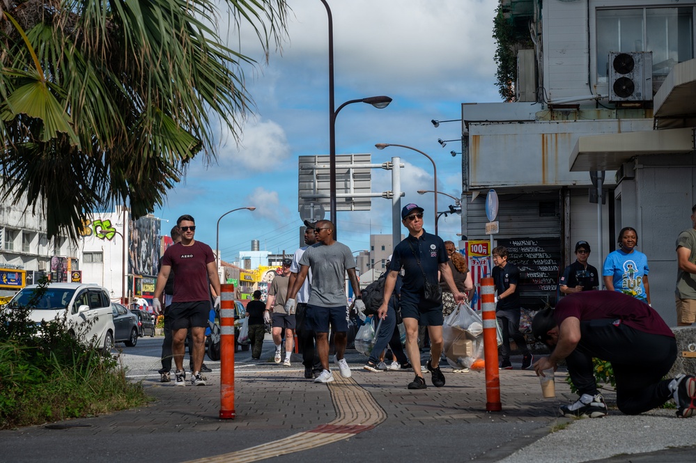 A group of people are walking along a urban sidewalk in what appears to be a Japanese city, likely Okinawa, based on the Japanese signage and architectural style visible. Several individuals are carrying bags, suggesting a community cleanup or volunteer activity. The scene is set on a busy commercial street with palm trees, storefronts, and street infrastructure visible.