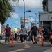 A group of people are walking along a urban sidewalk in what appears to be a Japanese city, likely Okinawa, based on the Japanese signage and architectural style visible. Several individuals are carrying bags, suggesting a community cleanup or volunteer activity. The scene is set on a busy commercial street with palm trees, storefronts, and street infrastructure visible.