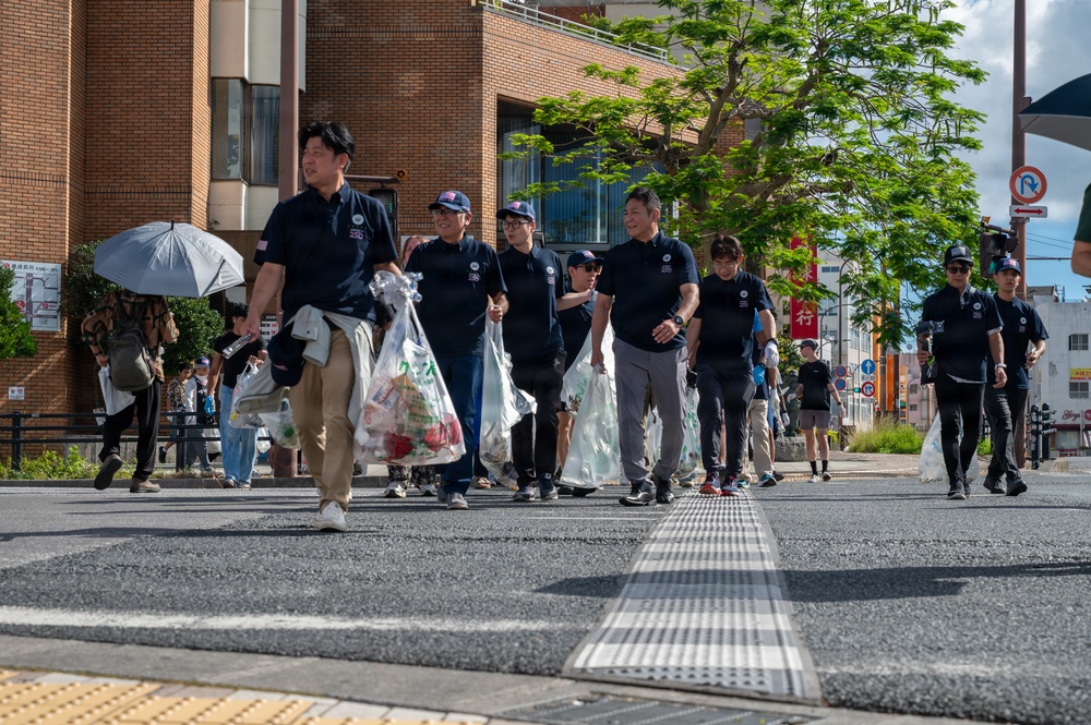 A group of people wearing matching dark navy polo shirts are walking across a street in what appears to be a Japanese urban area, carrying large plastic bags filled with collected trash or recyclables. The scene suggests an organized community cleanup event, with participants moving together as a coordinated group. The setting includes Japanese signage, brick buildings, and a large green tree in the background.