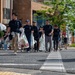 A group of people wearing matching dark navy polo shirts are walking across a street in what appears to be a Japanese urban area, carrying large plastic bags filled with collected trash or recyclables. The scene suggests an organized community cleanup event, with participants moving together as a coordinated group. The setting includes Japanese signage, brick buildings, and a large green tree in the background.