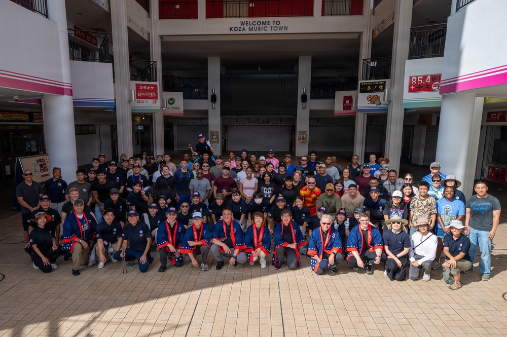 A large group of approximately 80-100 people pose for a group photo at Koza Music Town, a cultural venue in Okinawa, Japan. Several participants in the front rows are wearing matching dark blue happi coats (traditional Japanese festival jackets) with red and white accents, suggesting a cultural exchange or community event. The setting is an open-air atrium of the venue with Japanese and English signage visible.