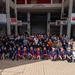 A large group of approximately 80-100 people pose for a group photo at Koza Music Town, a cultural venue in Okinawa, Japan. Several participants in the front rows are wearing matching dark blue happi coats (traditional Japanese festival jackets) with red and white accents, suggesting a cultural exchange or community event. The setting is an open-air atrium of the venue with Japanese and English signage visible.