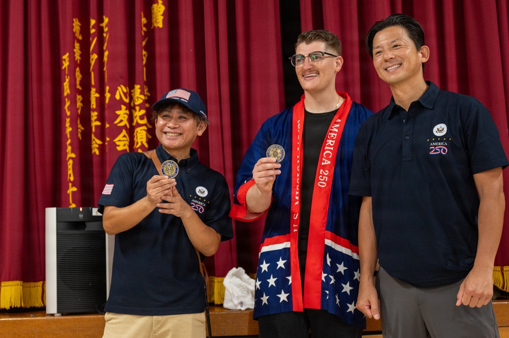 Three people are posing together on a stage in front of a red curtain with Chinese/Japanese characters. The central figure is wearing a patriotic American-themed robe and an 'America 250' sash, while all three individuals wear navy blue 'America 250' polo shirts. Two of the individuals are holding up commemorative coins and smiling for the photo.