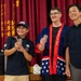 Three people are posing together on a stage in front of a red curtain with Chinese/Japanese characters. The central figure is wearing a patriotic American-themed robe and an 'America 250' sash, while all three individuals wear navy blue 'America 250' polo shirts. Two of the individuals are holding up commemorative coins and smiling for the photo.
