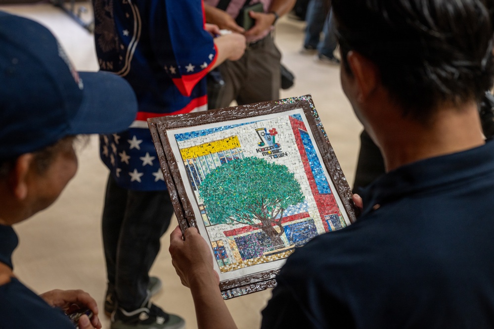 Two people are examining a framed mosaic artwork depicting a building scene with a prominent green tree in the foreground. The mosaic appears to reference 'Kazaa Music Town' based on visible text and logo on the artwork. The scene takes place in what appears to be an indoor gathering or event space with other people visible in the background.