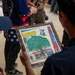 Two people are examining a framed mosaic artwork depicting a building scene with a prominent green tree in the foreground. The mosaic appears to reference 'Kazaa Music Town' based on visible text and logo on the artwork. The scene takes place in what appears to be an indoor gathering or event space with other people visible in the background.