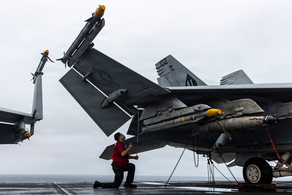 A naval aviation technician in a red shirt kneels on a carrier flight deck performing an inspection or maintenance check on an F/A-18 Hornet or Super Hornet. The aircraft has its wings folded and is loaded with multiple air-to-air missiles, secured to the deck with chains. The overcast sky and wet deck surface suggest at-sea operations in poor weather conditions.