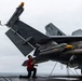 A naval aviation technician in a red shirt kneels on a carrier flight deck performing an inspection or maintenance check on an F/A-18 Hornet or Super Hornet. The aircraft has its wings folded and is loaded with multiple air-to-air missiles, secured to the deck with chains. The overcast sky and wet deck surface suggest at-sea operations in poor weather conditions.