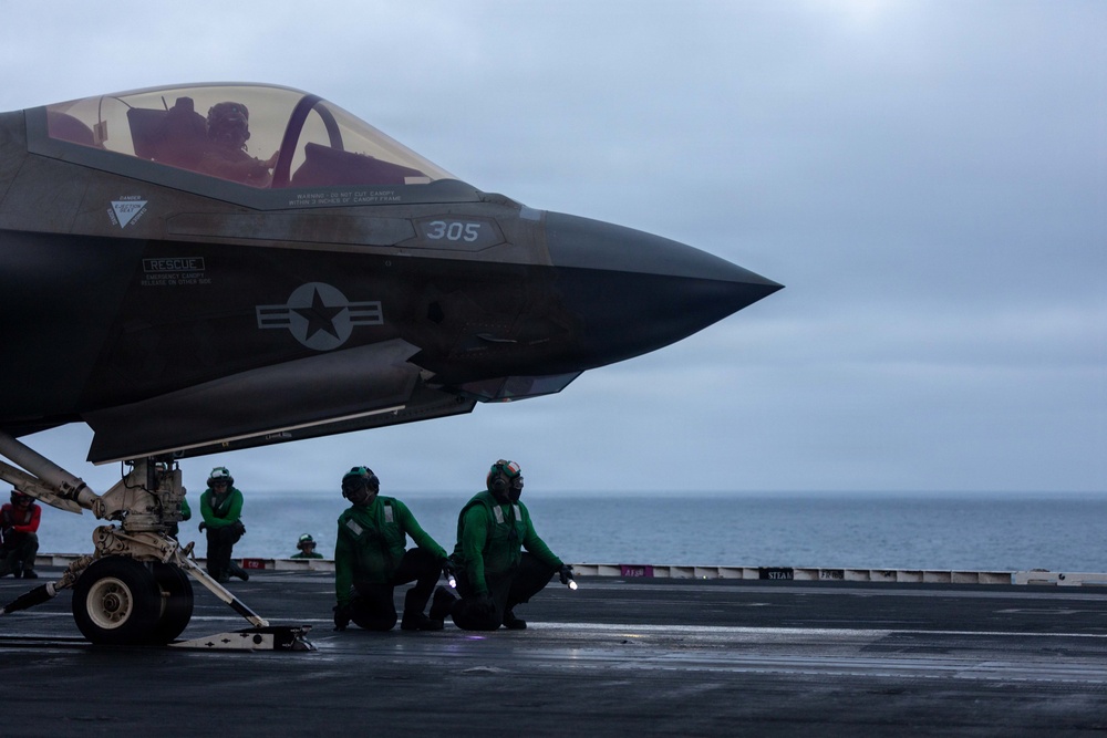 A U.S. military F-35 stealth fighter jet, numbered 305, is positioned on the wet flight deck of an aircraft carrier at sea under overcast skies. Several deck crew members dressed in green flight deck jerseys are kneeling and crouching near the aircraft, performing pre-launch or inspection checks. A pilot is visible in the cockpit, suggesting the aircraft is being prepared for launch operations.