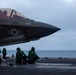 A U.S. military F-35 stealth fighter jet, numbered 305, is positioned on the wet flight deck of an aircraft carrier at sea under overcast skies. Several deck crew members dressed in green flight deck jerseys are kneeling and crouching near the aircraft, performing pre-launch or inspection checks. A pilot is visible in the cockpit, suggesting the aircraft is being prepared for launch operations.