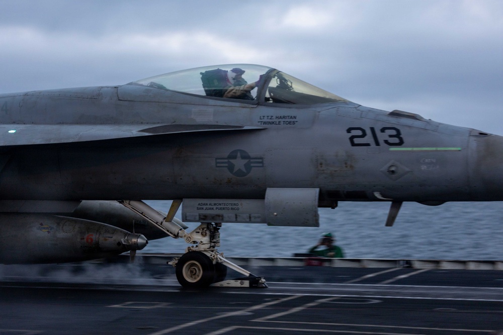 A U.S. Navy carrier-based jet aircraft numbered '213' is captured during a catapult launch from an aircraft carrier flight deck. The aircraft bears visible markings including a pilot name 'LT T.Z. HARITAN' with callsign 'TWINKLE TOES' and a crew chief dedication to 'ADAN ALMONTEOTERO, SAN JUAN, PUERTO RICO'. A flight deck crew member in a green jersey is visible in the background.