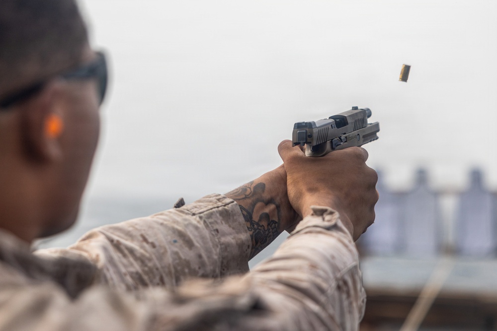 A military or law enforcement personnel in desert camouflage uniform is firing a semi-automatic pistol at a shooting range. A brass cartridge casing is visibly ejecting from the firearm, captured mid-air to the upper right. Blurred target silhouettes are visible in the background, confirming a controlled range environment.