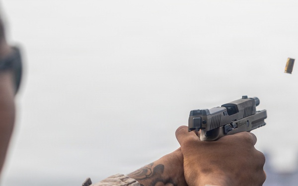A military or law enforcement personnel in desert camouflage uniform is firing a semi-automatic pistol at a shooting range. A brass cartridge casing is visibly ejecting from the firearm, captured mid-air to the upper right. Blurred target silhouettes are visible in the background, confirming a controlled range environment.