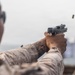 A military or law enforcement personnel in desert camouflage uniform is firing a semi-automatic pistol at a shooting range. A brass cartridge casing is visibly ejecting from the firearm, captured mid-air to the upper right. Blurred target silhouettes are visible in the background, confirming a controlled range environment.