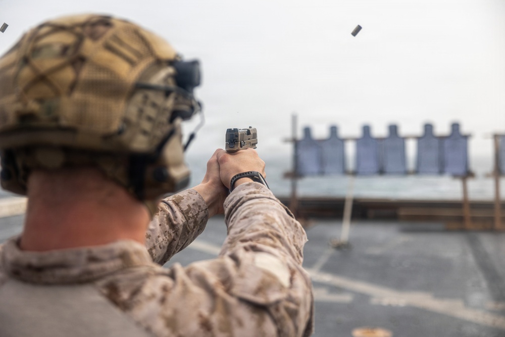 A military service member in desert camouflage uniform and tactical helmet is conducting pistol qualification or training on what appears to be a ship's deck. The shooter is firing a semi-automatic pistol at a row of silhouette targets downrange, with a spent brass casing visibly ejecting from the firearm. The setting and uniform suggest a U.S. Marine Corps marksmanship exercise aboard a naval vessel.