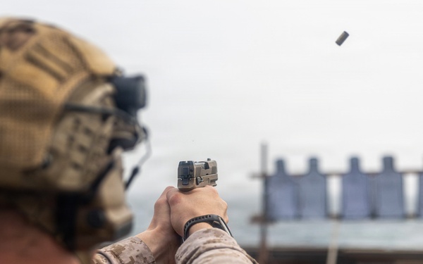 A military service member in desert camouflage uniform and tactical helmet is conducting pistol qualification or training on what appears to be a ship's deck. The shooter is firing a semi-automatic pistol at a row of silhouette targets downrange, with a spent brass casing visibly ejecting from the firearm. The setting and uniform suggest a U.S. Marine Corps marksmanship exercise aboard a naval vessel.