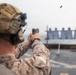 A military service member in desert camouflage uniform and tactical helmet is conducting pistol qualification or training on what appears to be a ship's deck. The shooter is firing a semi-automatic pistol at a row of silhouette targets downrange, with a spent brass casing visibly ejecting from the firearm. The setting and uniform suggest a U.S. Marine Corps marksmanship exercise aboard a naval vessel.