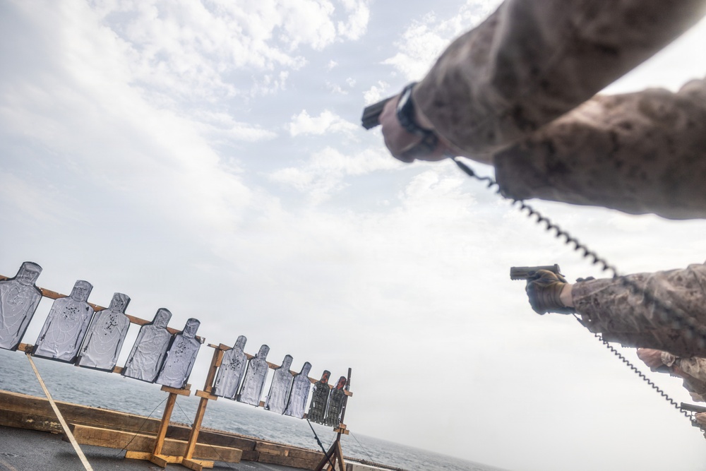 Military personnel in camouflage uniforms are conducting a pistol qualification or training exercise on what appears to be the deck of a naval vessel. Multiple human-silhouette paper targets are mounted on wooden frames along the ship's railing, with visible bullet holes indicating active shooting. The image is taken from a low, wide-angle perspective, creating a dramatic composition.