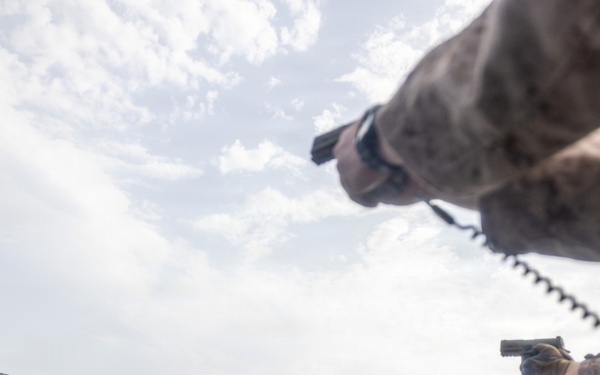 Military personnel in camouflage uniforms are conducting a pistol qualification or training exercise on what appears to be the deck of a naval vessel. Multiple human-silhouette paper targets are mounted on wooden frames along the ship's railing, with visible bullet holes indicating active shooting. The image is taken from a low, wide-angle perspective, creating a dramatic composition.