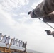 Military personnel in camouflage uniforms are conducting a pistol qualification or training exercise on what appears to be the deck of a naval vessel. Multiple human-silhouette paper targets are mounted on wooden frames along the ship's railing, with visible bullet holes indicating active shooting. The image is taken from a low, wide-angle perspective, creating a dramatic composition.